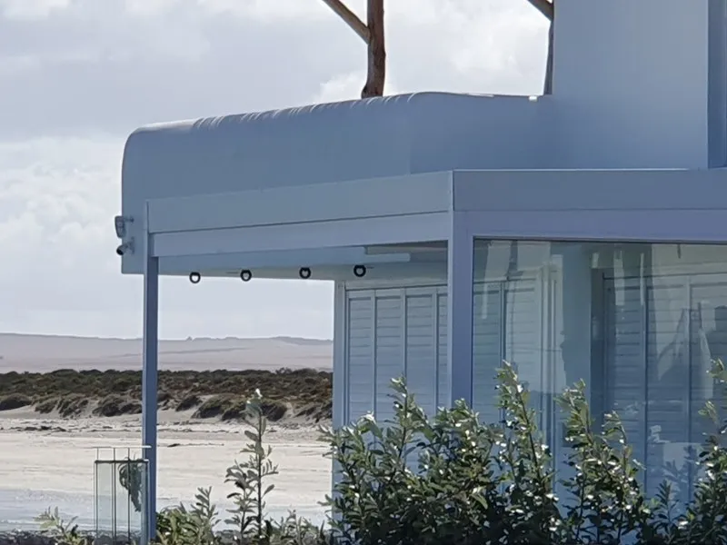Pale grey curved aluminium canopy with recessed downlights over the glazed corner of a beach house, dunes and sand behind
