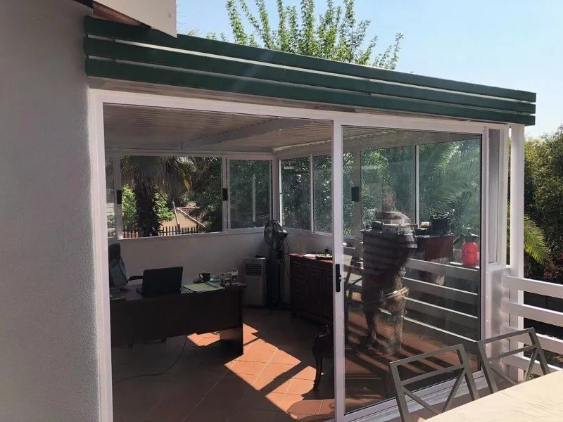 Aluminium cover with a green fascia over a patio closed in with sliding glass panels, garden beyond