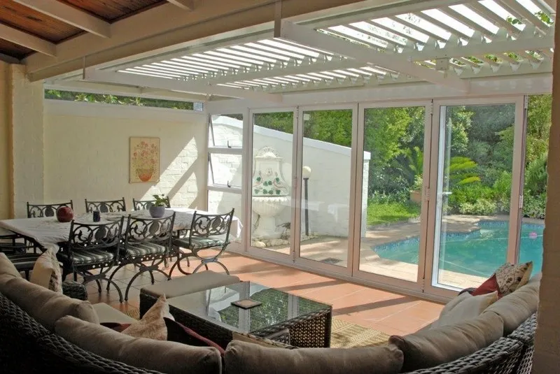 White louvre roof over an enclosed patio room with folding glass doors, cast-aluminium dining set and a pool beyond