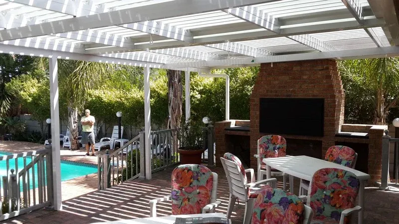 White louvre pergola over a poolside entertainment area with a face-brick braai and a wall-mounted television