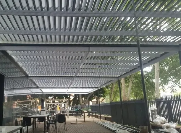 Large charcoal louvre roof over a paved courtyard of tables, mature trees overhead